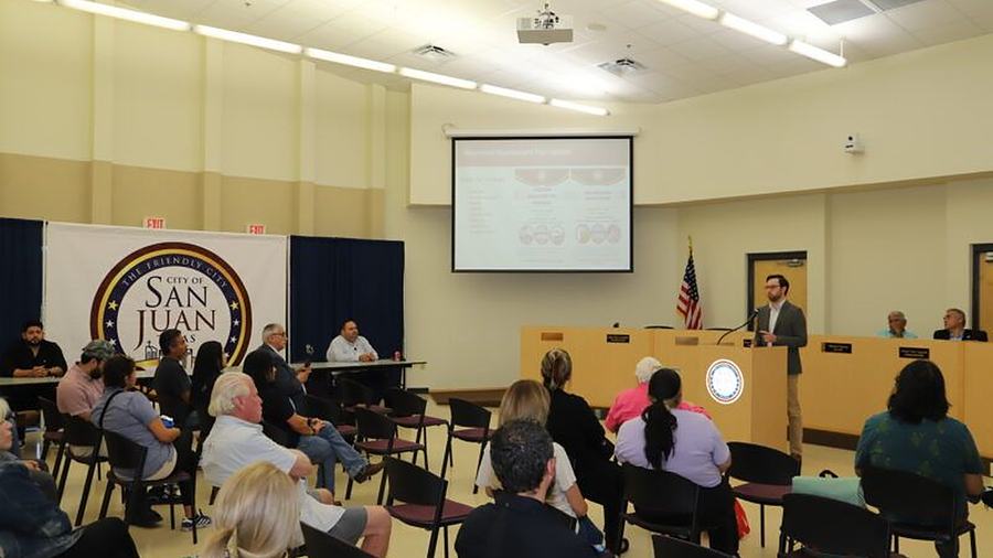 The San Juan City Commission in session in the council chamber, the city's seal on a banner behind the dais and residents seated facing them.