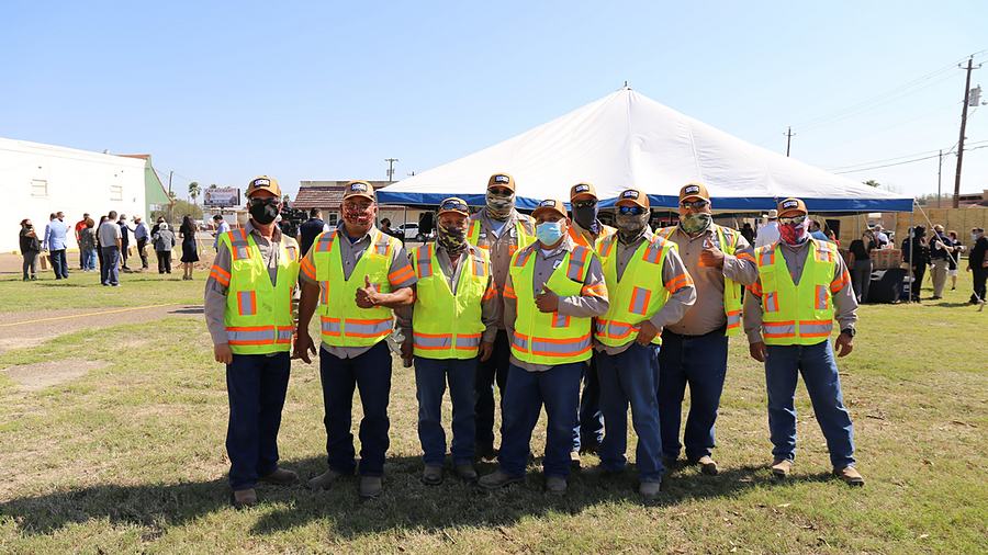 A City of San Juan crew standing together outdoors in high-visibility vests and caps, a marquee and a gathering behind them.
